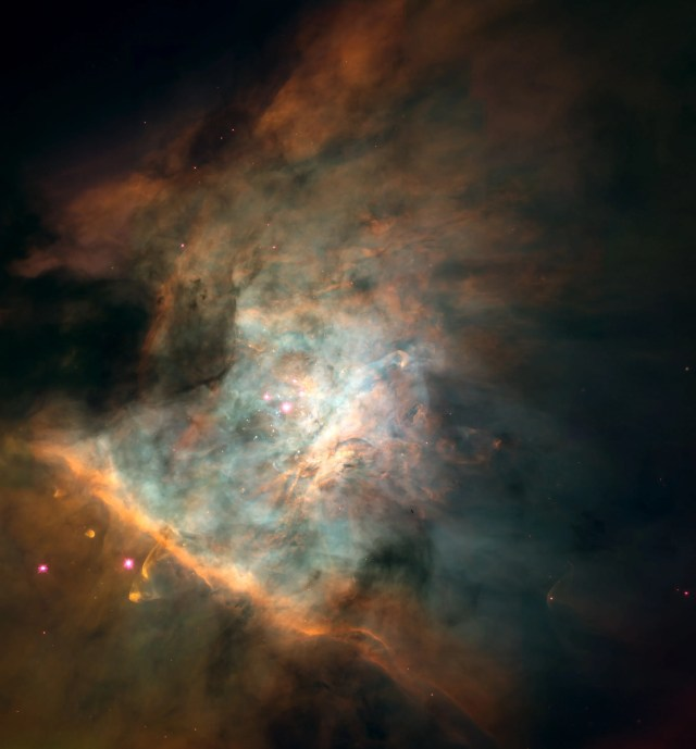 Dark clouds ring the image. The middle of the image holds bright clouds in colors of green, light blue, orange, yellow and white. Near the image center is a cluster of stars called the Trapezium.