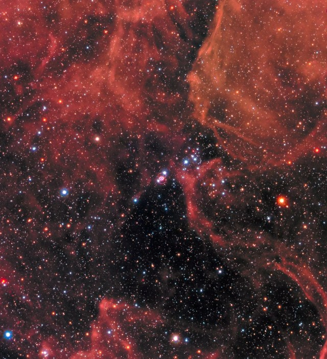 Black background filled with stars. Bright-red clouds fill the upper half of the image. Supernova 1987a is at image center. It is a bright point of light surrounded by a bright, pink-white ring. two red rings circle the central ring and form a figure eight. Two bright stars appear like diamonds. One along the upper ring, and the other along the lower red rings.