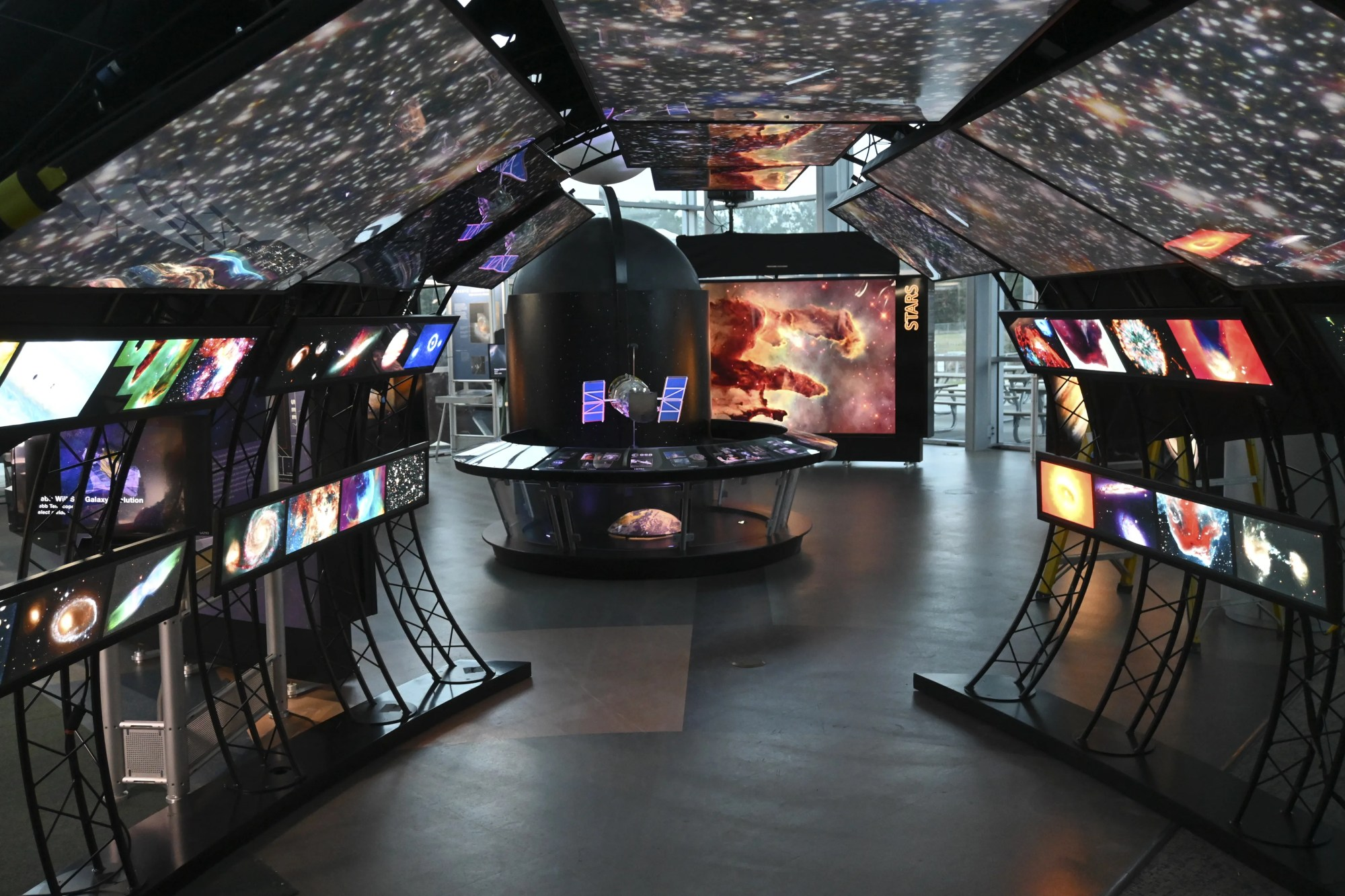 View of from within the tunnel of the Hubble exhibit. Illuminated Hubble images of galaxies and stars overhead, individual objects along the walls. Hubble Space Telescope model at the end of of the tunnel. Large illuminated Eagle Nebula image beyond.
