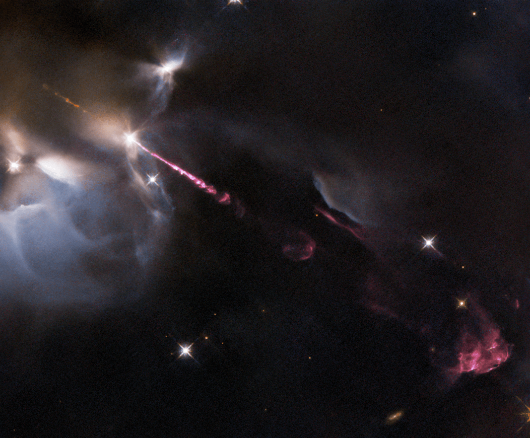 Dark cloud of gas and dust a with bright-white glowing outline in the upper left. pinkish-red jet of material extends from the cloud to the lower right where billowing reddish-pink cloud forms. background stars
