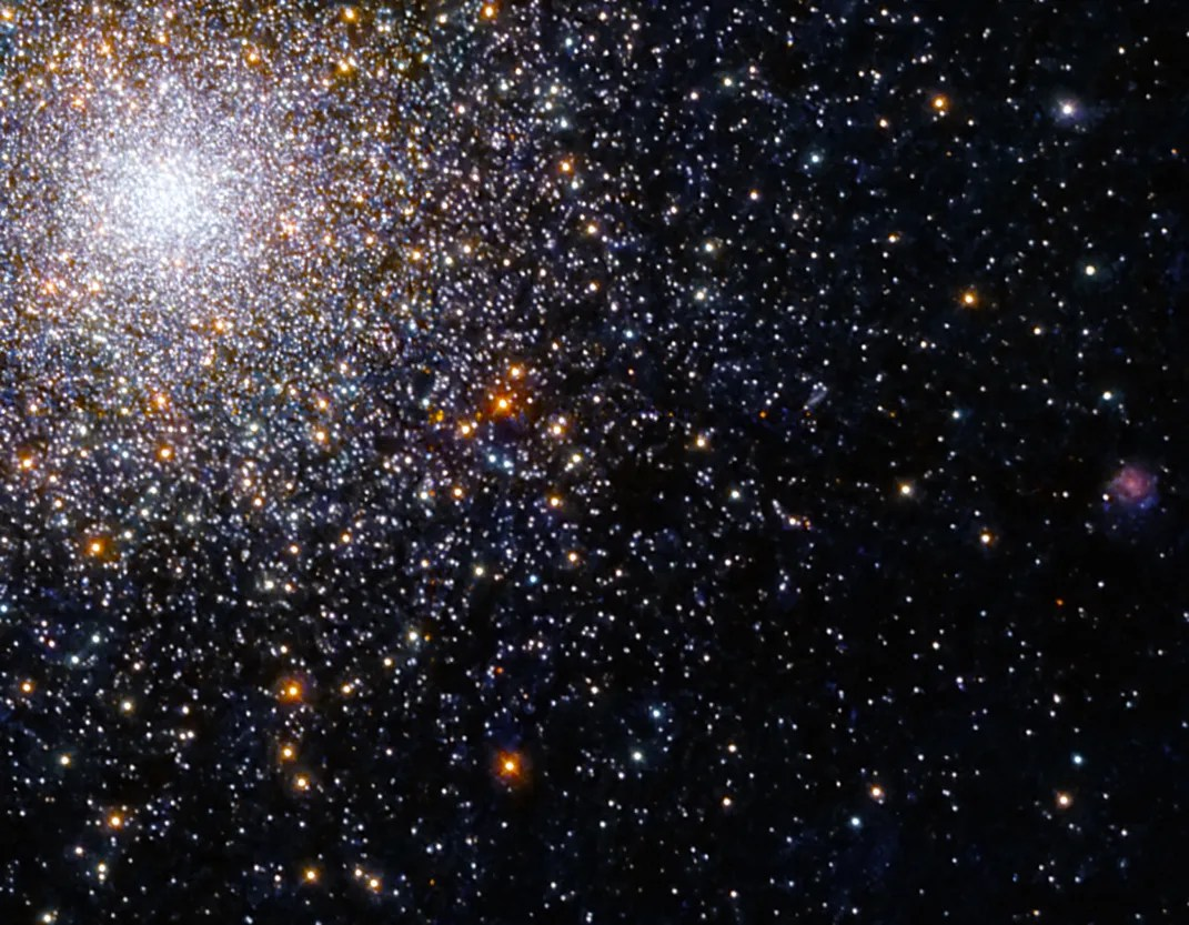 Bright-white spherical cluster of stars in the upper left corner. Stars taper off down and out  to the right of the cluster against a black background. Reddish-orange stars dot the scene.