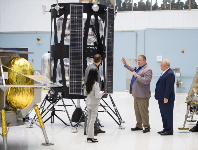 Four people gather around in front of the Research and Development of Intuitive Machines’ lunar lander. The man second from the right, Time Crain, speaks while his hand is raised towards the lander to the others around him. Thomas Zurbuchen, second from the left, looks at the lunar lander that is twice his size while Crain is talking to the group. The lander has a black outer shell with silver structures that are resting within.