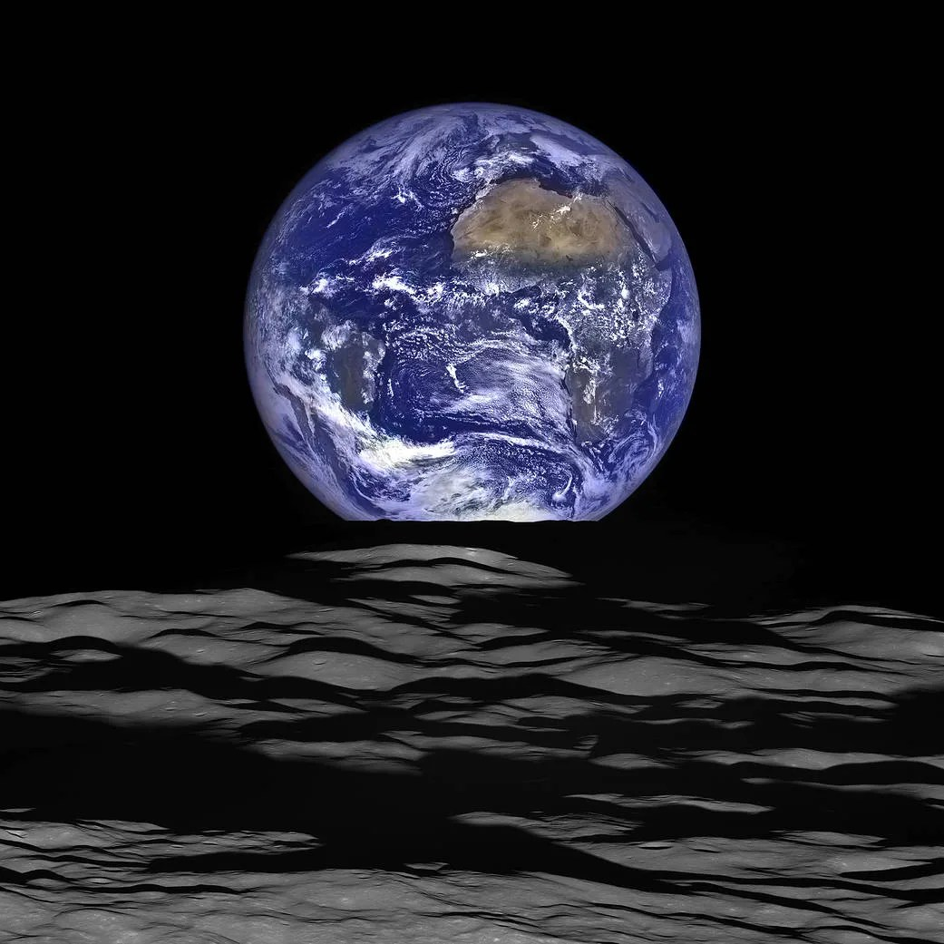 The brightly illuminated Earth in nearly full view over the dimly lit lunar surface in the foreground. The Earth is seen in the center but is partly cut off on the bottom by the Moon’s surface. There is a flurry of white clouds in the southern hemisphere past the tip of Africa. The African continent peaks through Earth’s busy atmosphere and the sapphire blue ocean shines brilliantly around it.