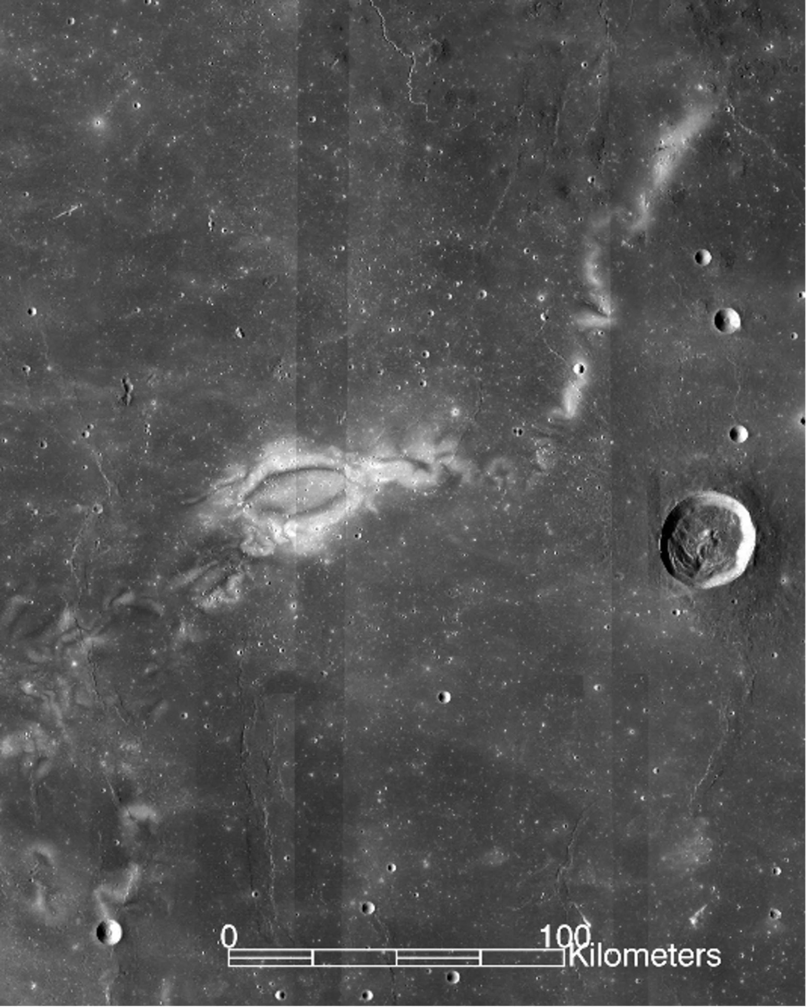 The image is of a large geological feature on the lunar surface. The majority of the image is dark grey and appearing to show a relatively flat expanse. A few small craters are scattered throughout and one large one on the far right. In the center-left of the image, Reiner Gamma appears as a light-colored oval with wispy, white edges like a cloud. Additional scattered streaks of white extend up and to the right of the swirl. Along the bottom of the image is a scale indicating that the main part of the swirl