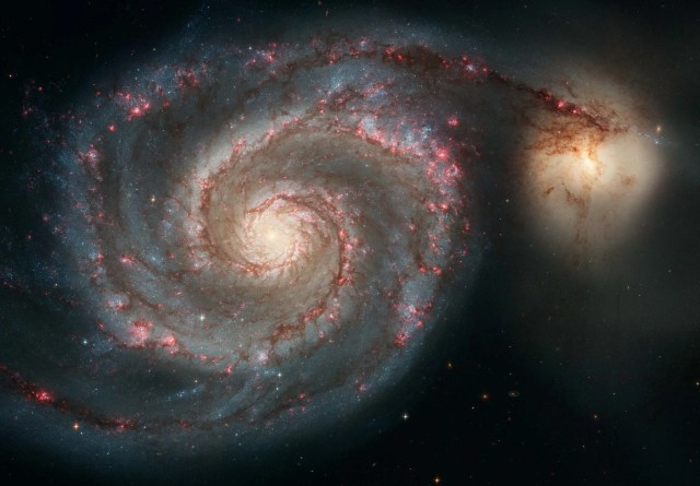 A spiral galaxy seen from overhead, with its two major arms spiraling out from its bright white core. At the end of the arm on the right is another bright white blob, this is a second galaxy. The arms are bluish and purple, peppered with countless stars.