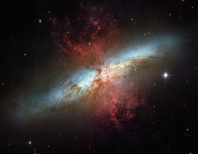 A white band of stars that cuts across a black background, from the lower left to the upper right, is the galaxy M82. Reddish brown gas and dust overlays the galaxy concentrated in the center of the image and fanning out above and below the white band of the galaxy. Black background is dotted with stars.