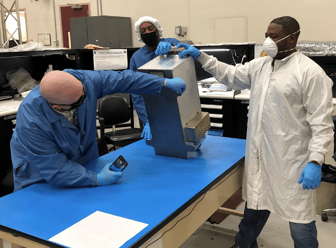 A metal mechanical box sits at an angle on a table, as three individuals - wearing gloves, lab coats and face masks - examine the box by touching the surface and photographing the box.
