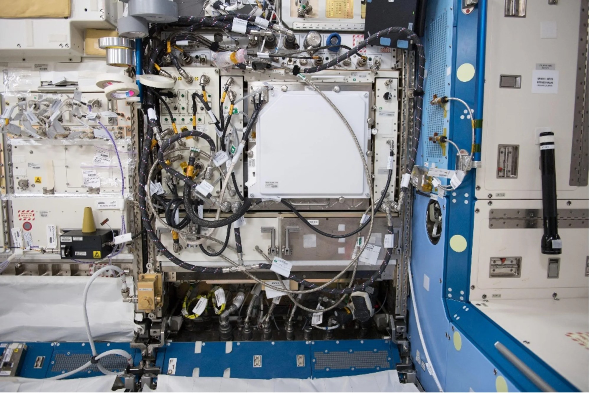 A complex metal box is set into the inside wall of the space station. Black and silver wires and tubes crisscross the front surface of the box, connecting the environmental control system in the left half of the box to the plant growth chamber in the right half.