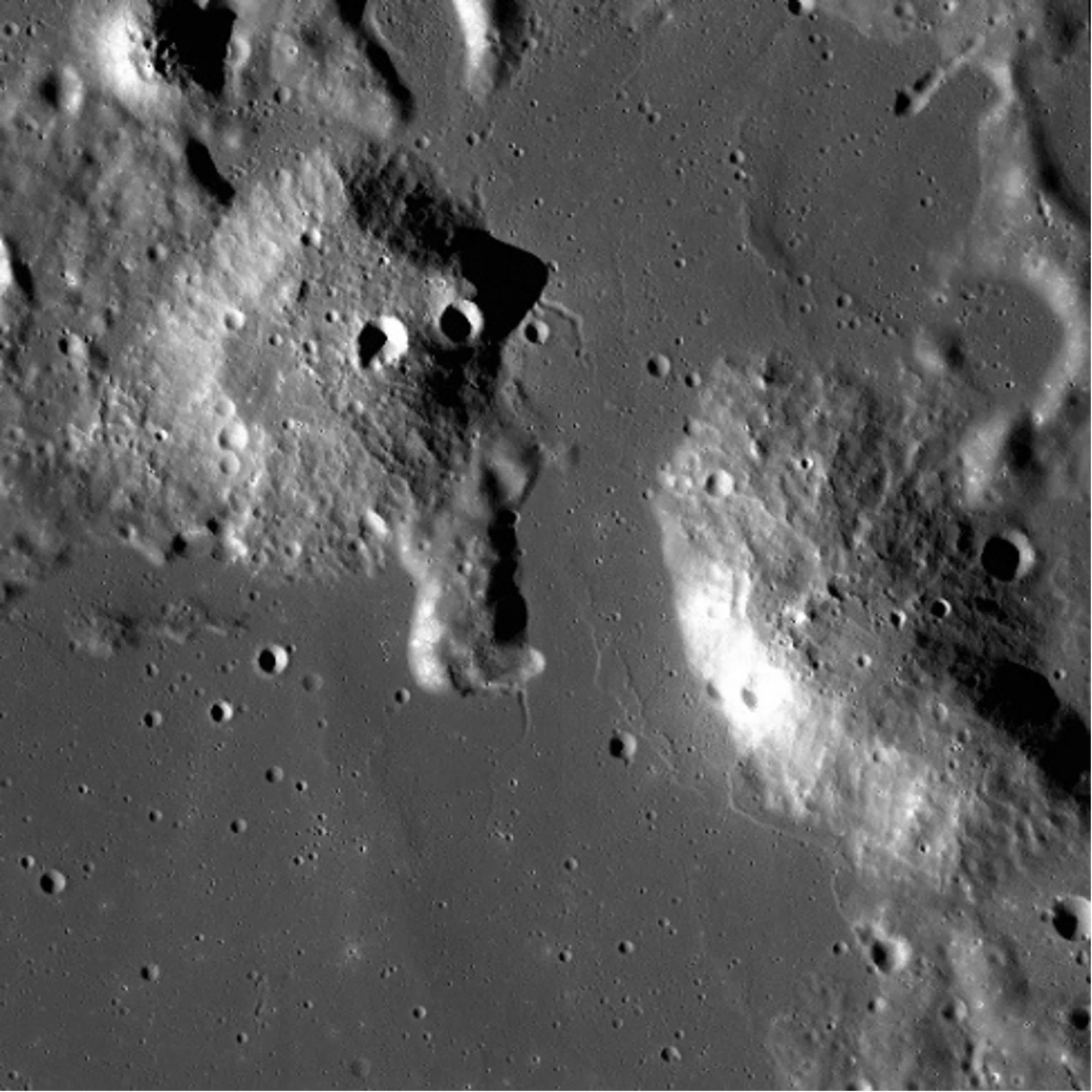 Pictured is an image of the lunar surface. The surface is relatively flat apart from two large mounds and some small, scattered craters. One sits in the bottom right corner and the other is towards the top left.  The top left mound has two prominent craters that are located on the middle-left side of the mound.