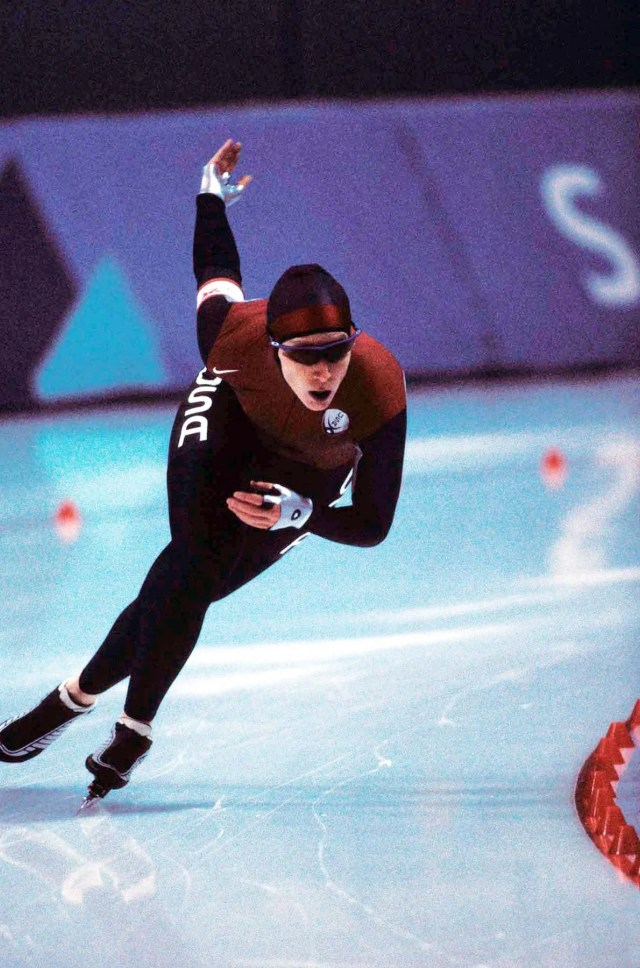 A skater wearing a red and black uniform with USA emblazoned on it in white letters skates across a rink. She wears googles and a close-fitting black hat.