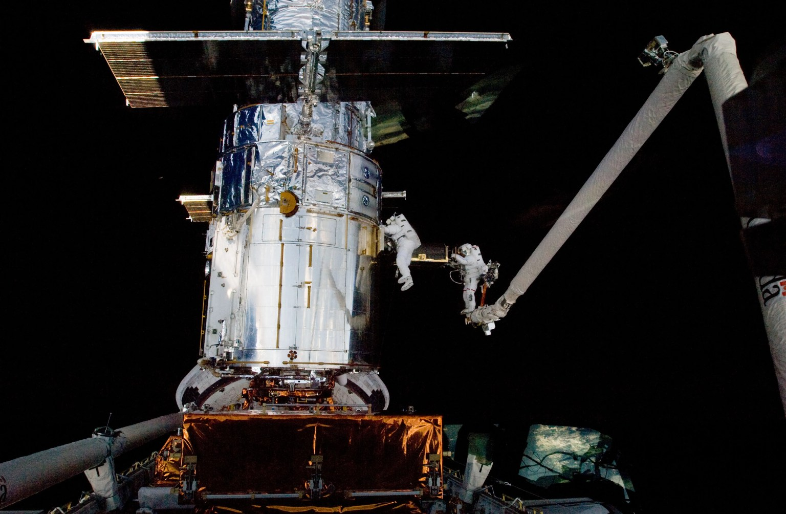 Two astronauts in white space suits tethered on an arm servicing the Hubble Space Telescope in space.
