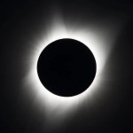 Against a black background, the Sun shines out around a black circle. The Sun appears as wispy white streams of light.