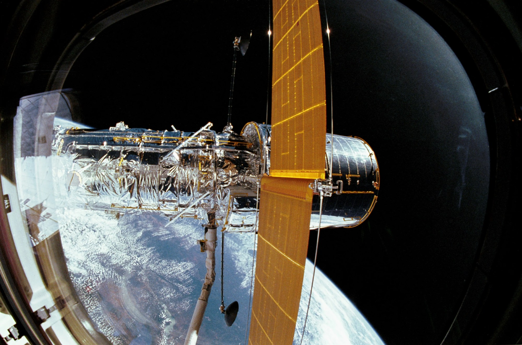 The Hubble Telescope in space gripped by the space shuttle's robotic arm. One solar panel is deployed. The round rim of the window through which the picture was taken is visible.