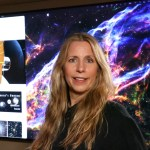 headshot of a woman with blonde hair in a black shirt standing in front of a hubble image on a tv monitor