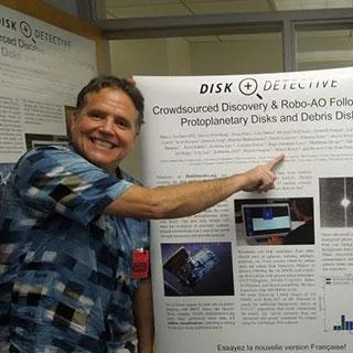 Photo of a smiling man wearing a short sleeve shirt and pointing at a large poster filled with text and universe imagery.