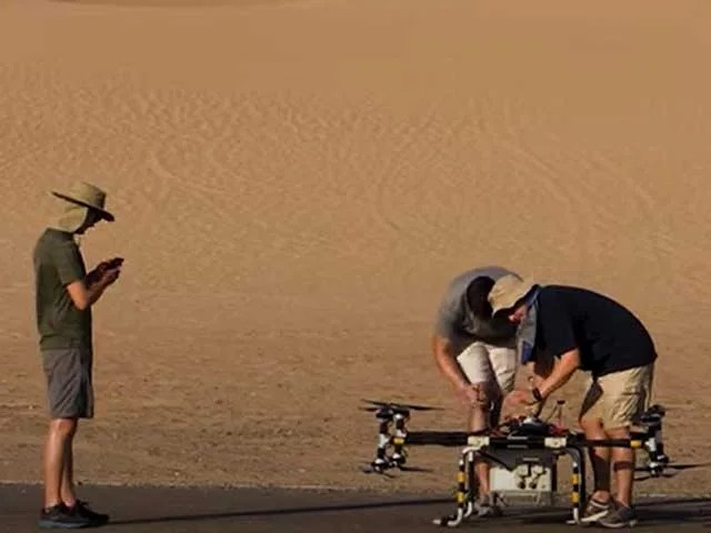 Two humans are bending over a model spacecraft. A third human is shown watching at the left. A sandy surface is shown in the background.