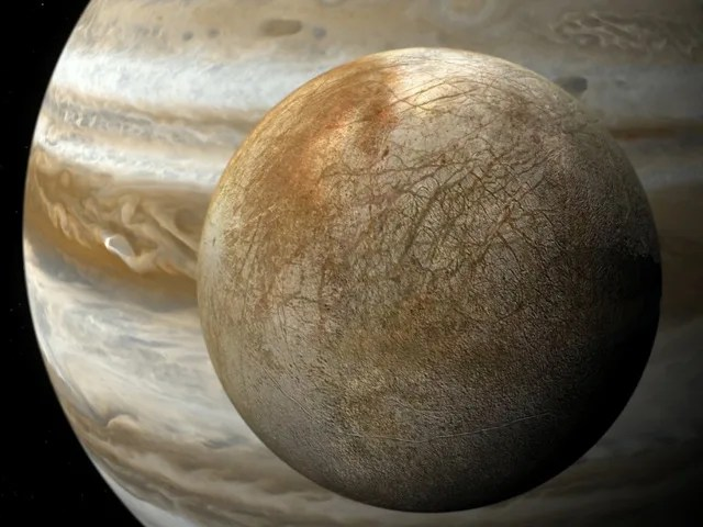 The brown and grey scratched surface of Europa is shown in front of the large planet of Jupiter.