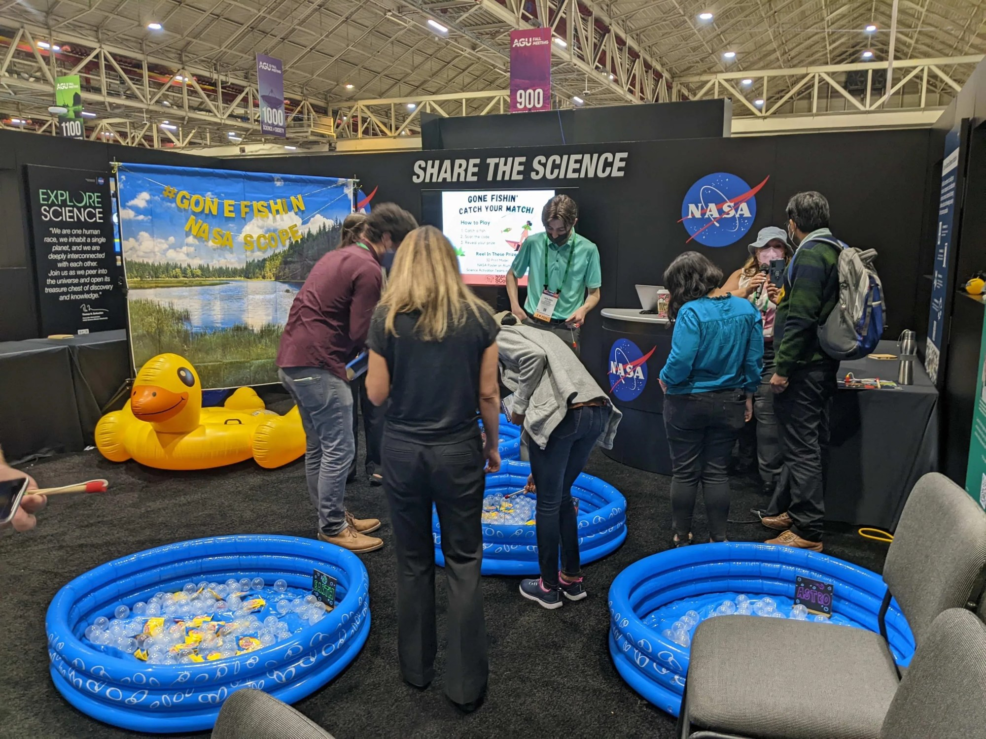 Learning area of the NASA booth showing inflatable swimming pools filled with fish, balls, etc. In the background is a backdrop of a lake with the title of Gone Fishin' NASA SCoPE and a large inflatable duck in front of it. Around the area, people are using fishing poles to catch fish, scan QR codes, and gathering giveaways.