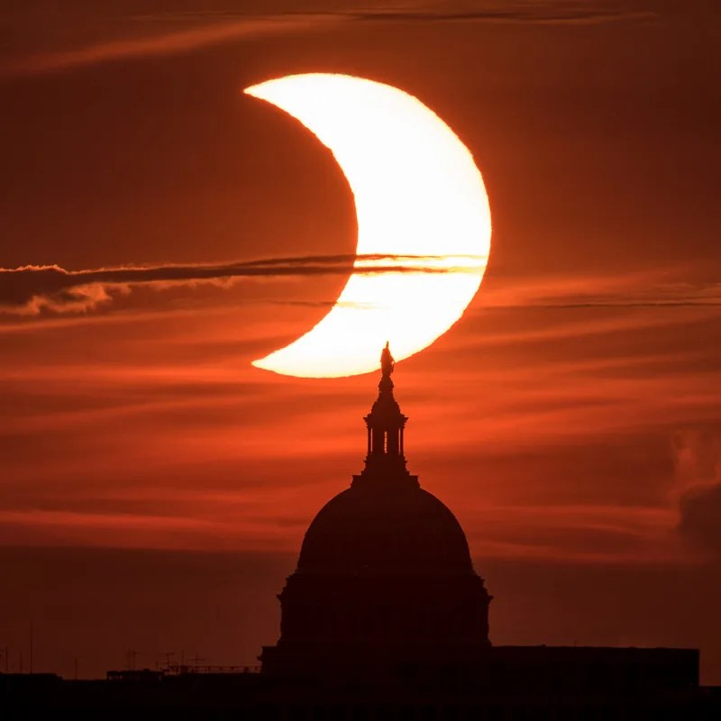 A partial solar eclipse is seen as the Sun rises behind the United States Capitol Building, Thursday, June 10, 2021, as seen from Arlington, Virginia. The sky is a dark red and some strands of clouds cross in front of the partial Sun.