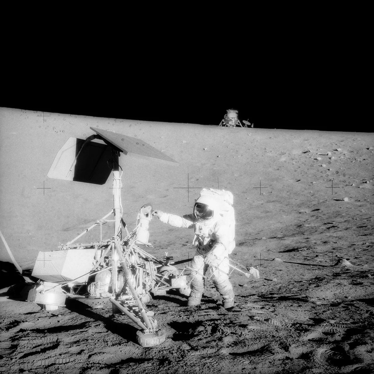 Astronaut in a white spacesuit standing on the surface of the Moon next to a robotic spacecraft with his human crew vehicle on the horizon.