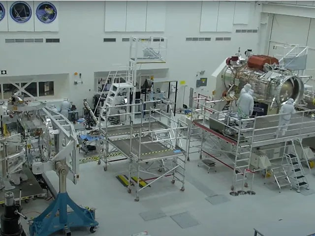 A room containing numerous platforms with ladders. People in white cleanroom suits are shown working on spacecraft equipment.