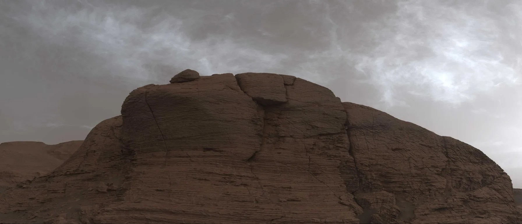 A bare orange-brown rocky cliff or outcropping with a rounded top fills most of the picture. Above it the sky is grey with a scattering of white, diffuse clouds.