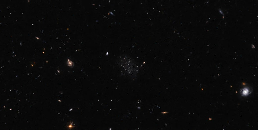 A black, mostly empty field with a variety of stars and galaxies spread across it. Most are very small. A couple of galaxies and stars are larger with visible details. In the centre is a relatively small, irregularly-shaped galaxy; it is formed of many very small stars and a few slightly larger, bright stars, all surrounded by a very faint glow that marks the borders of the galaxy.