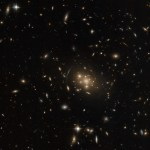 A cluster of large galaxies, surrounded by various stars and smaller galaxies on a dark background. The central cluster is mostly made of bright elliptical galaxies that are surrounded by a warm glow. Nearby the cluster is the stretched, distorted arc of a galaxy, gravitationally lensed by the cluster.