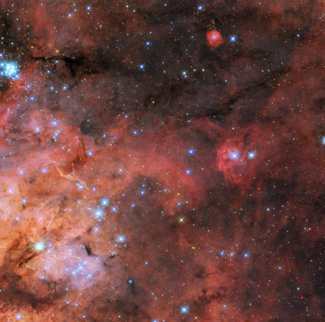 Wispy, nebulous clouds extend from the lower-left of the image. At the top and right the dark background of space can be seen through the sparse nebula. Along the left and in the corner are many layers of brightly-colored gas and dark, obscuring dust. A cluster of small, bright blue stars in the same corner expands out across the image. Many much smaller stars cover the background.