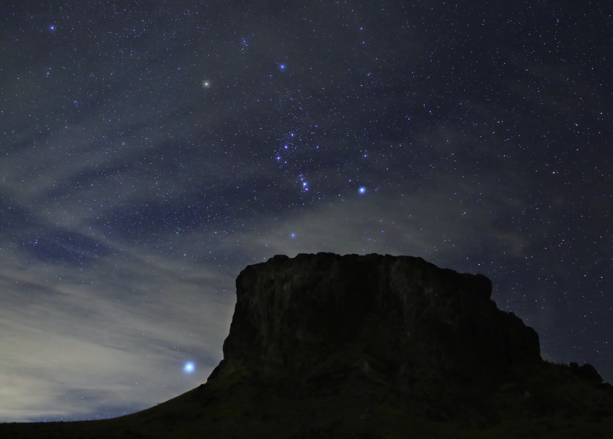 The bright stars in the constellation Orion appear above a rocky mesa at night. The bright star Sirius is seen to the left of the mesa.