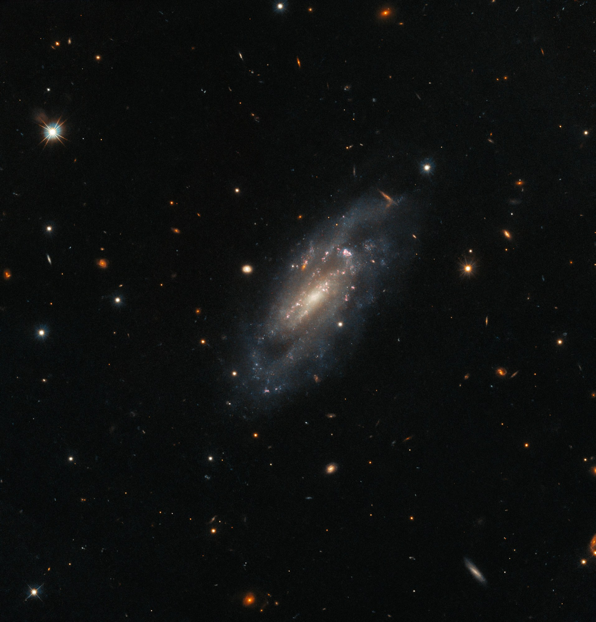 A spiral galaxy, a fuzzy oval tilted diagonally and partially towards the viewer. The center glows in warm colors, and has two prominent spiral arms around it, with bright points of star formation. The galaxy appears centrally in a field of small stars and galaxies on a dark background.