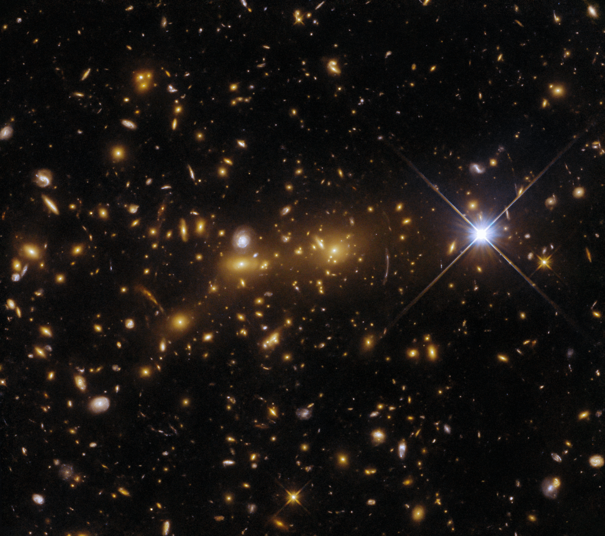 A cluster of elliptical galaxies, visible as a dense crowd of oval shapes, each glowing orange around a bright core. Various other galaxies are dotted all around, a few being small spirals. A bright star with four long spikes stands out at the right.