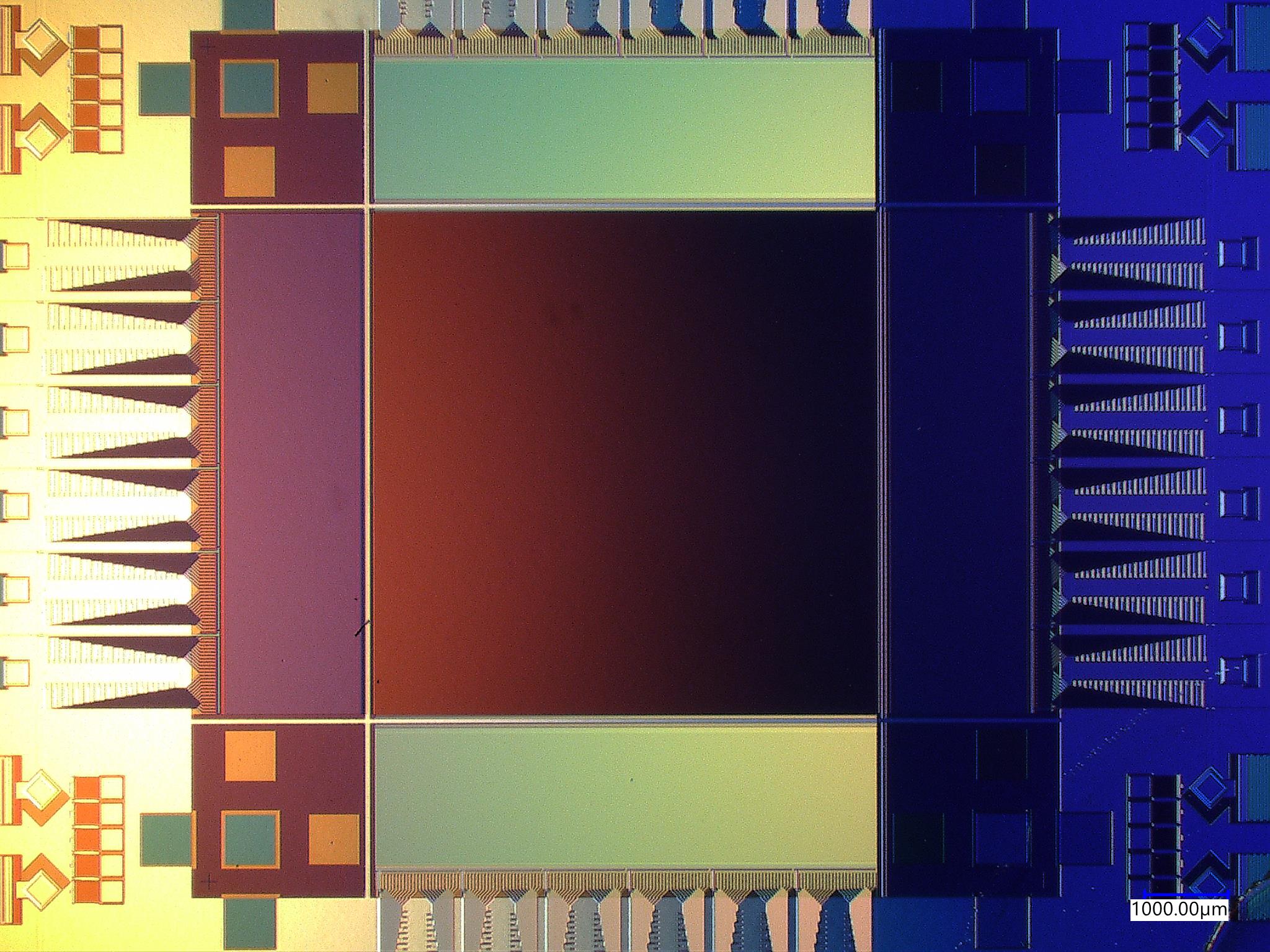 A microscope image of a rectangular chip, showing the different parts of the superconducting camera, including imaging area and ancillary electronics. The chip has a colorful patina, with hues of yellow on the left, red in the center, and blue on the right.