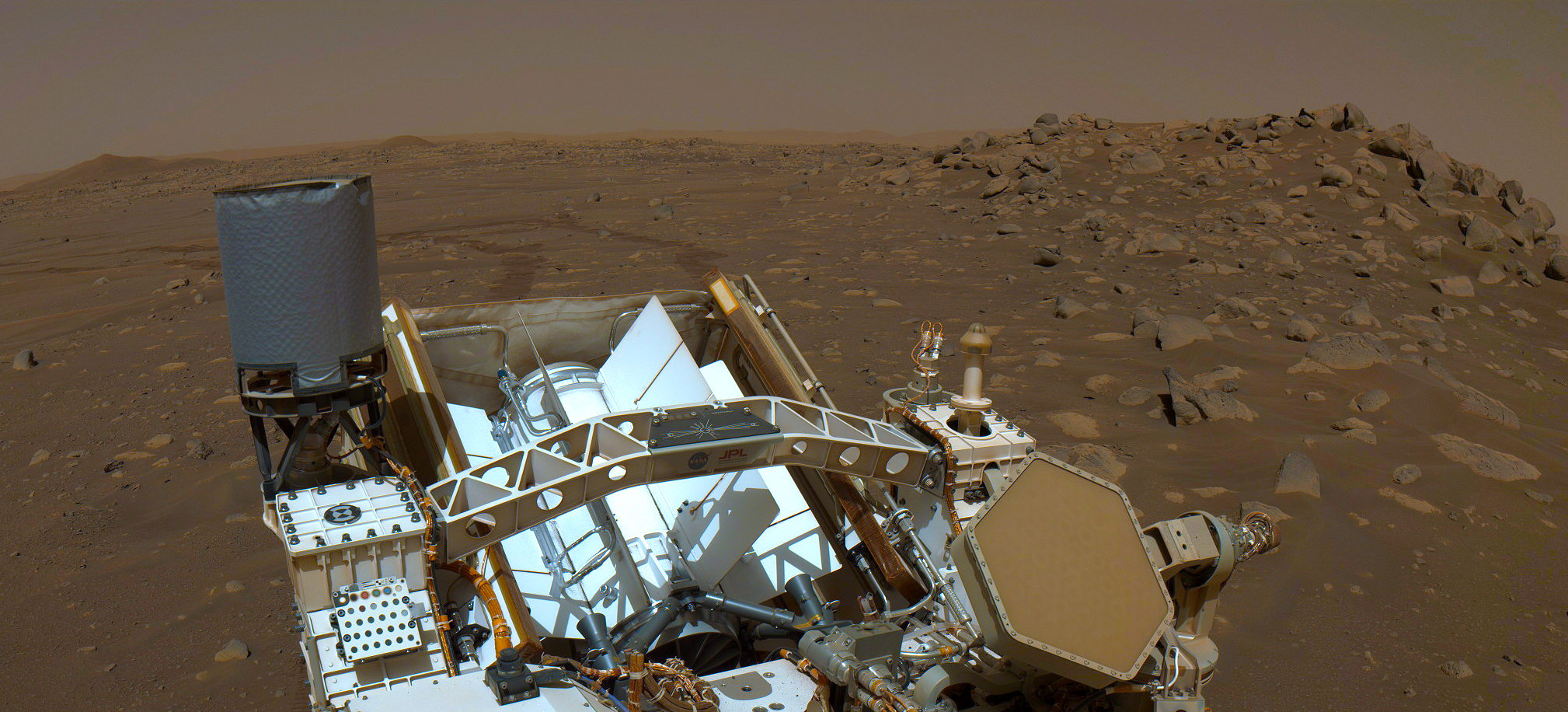 This view looking across the back of the Perseverance rover shows the rover's Radioisotope Thermoelectric Generator (RTG). A rocky, red landscape dotted with rocks fills the horizon.