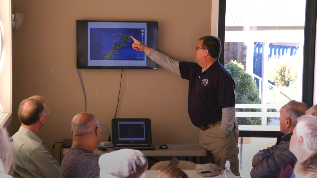 Man pointing at a map of New Zealand on a television monitor in front of a group of people.