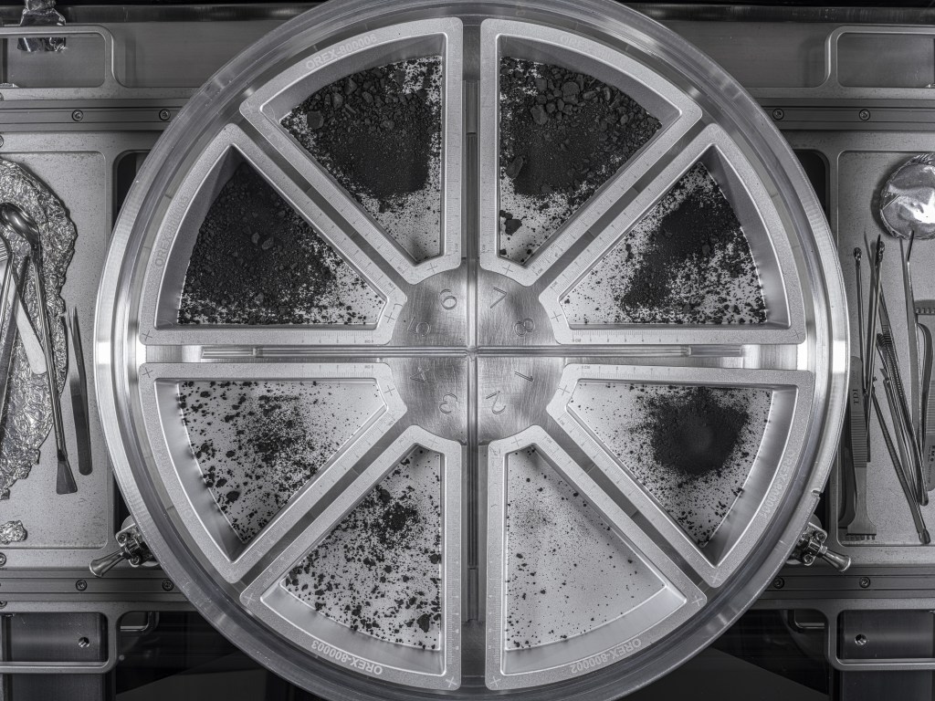 Silver metal wheel-shaped container with black-gray regolith inside the pie-shaped container areas