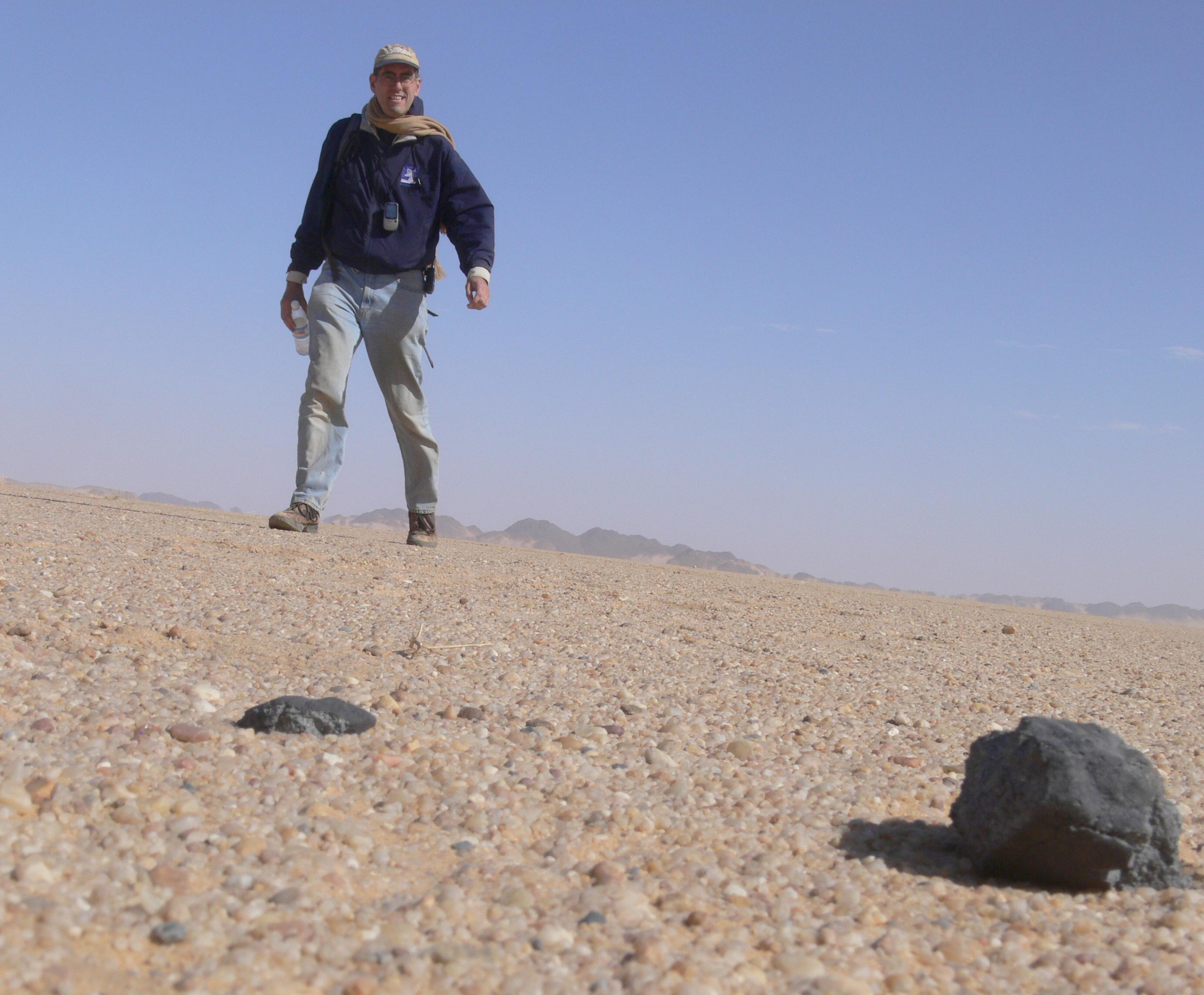 A man in a blue jacket, jeans, a tan scarfc and a baseball type cap walks toward a rock in the desert.
