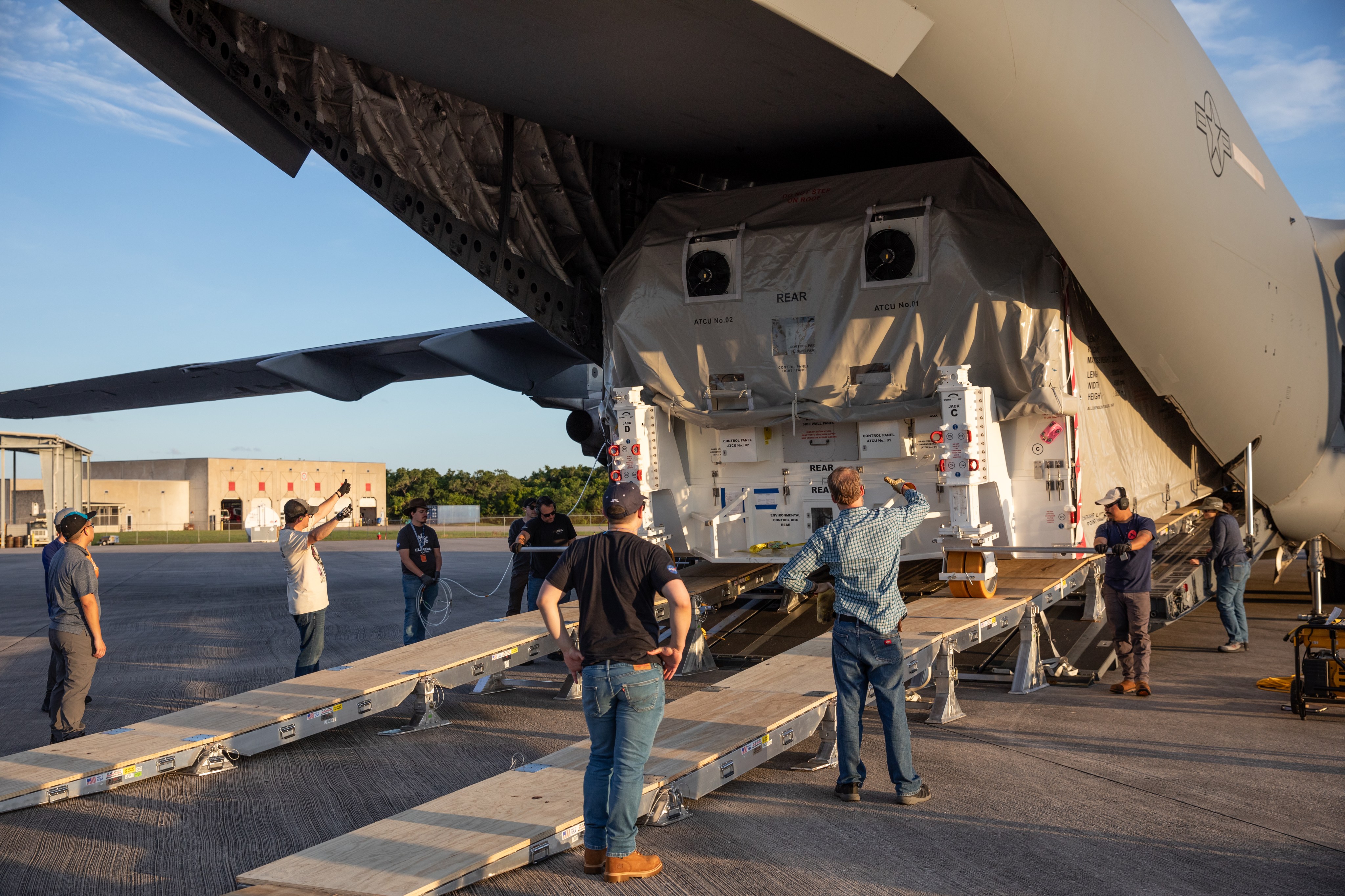 The back of a green transport aircraft is open like a giant duck's bill, and the shipping container hold Europa Clipper can be seen sitting inside. A ramp extends from the back of the plane. Workers are giving hand signals as they guide the shipping container off the plane.