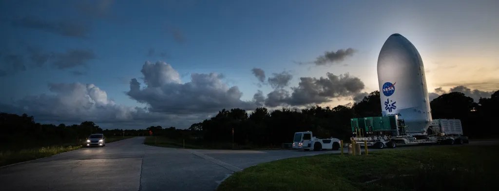 Europa Clipper, enclosed in its white, payload fairing, as it is moved from one building to another. Two trucks are nearby. The spacecraft towers over the escort vehicles.