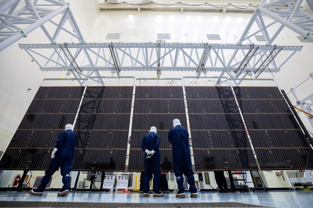 Three workers in blue protective jumpsuits with white head covers stand in front of part of the solar arrays for Europa Clipper.