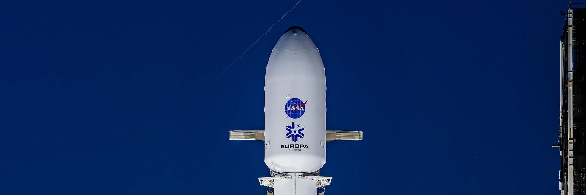 Europa Clipper sits atop a rocket at Launch Pad 39A at Kenned Space Center. The spacecraft is enclosed in a white fairing with the blue NASA logo, and the Europa Clipper mission identifier visible.