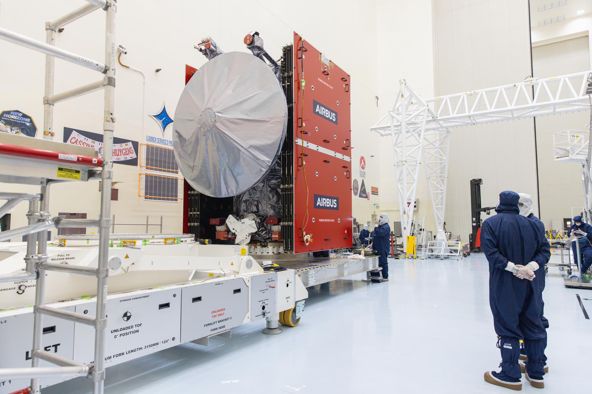 The Europa Clipper spacecraft sits on a large, wheeled work platform. It high silver high gain antenna is facing toward us. Its solar panels are folded up and covered in red protective shields with Airbus printed on them. Several workers in blue jumpsuits are nearby.