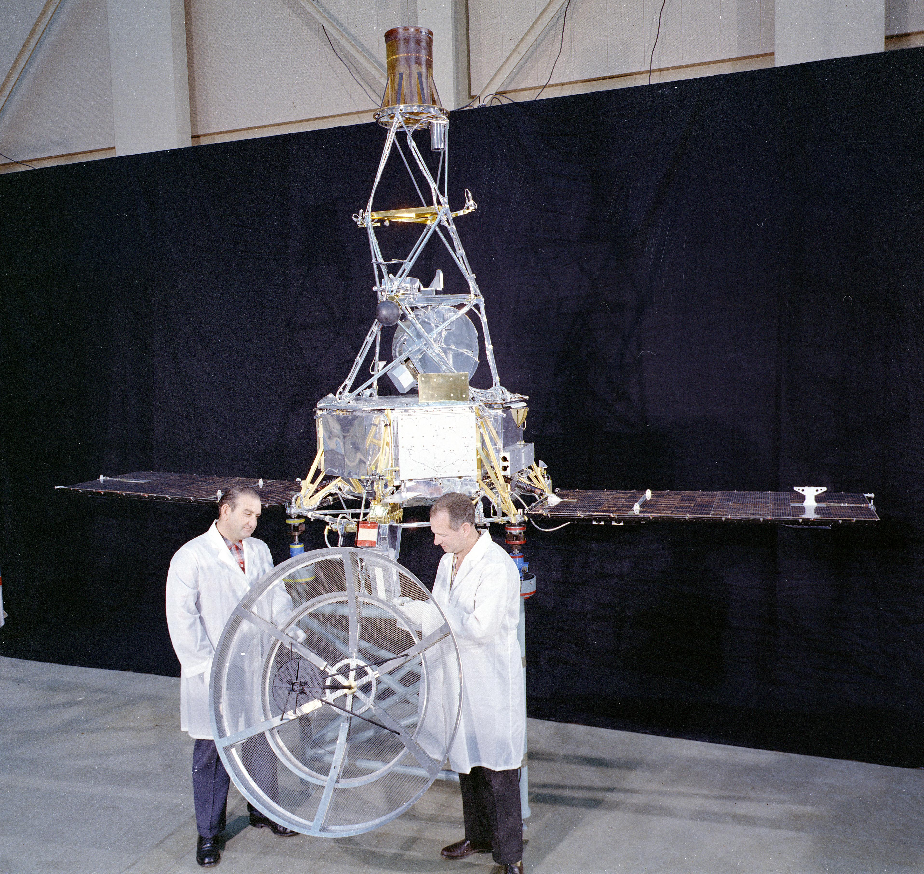 Two engineers in white lab boats inspect the Mariner 1 spacecraft. The spacecraft is about twice as tall as the men.