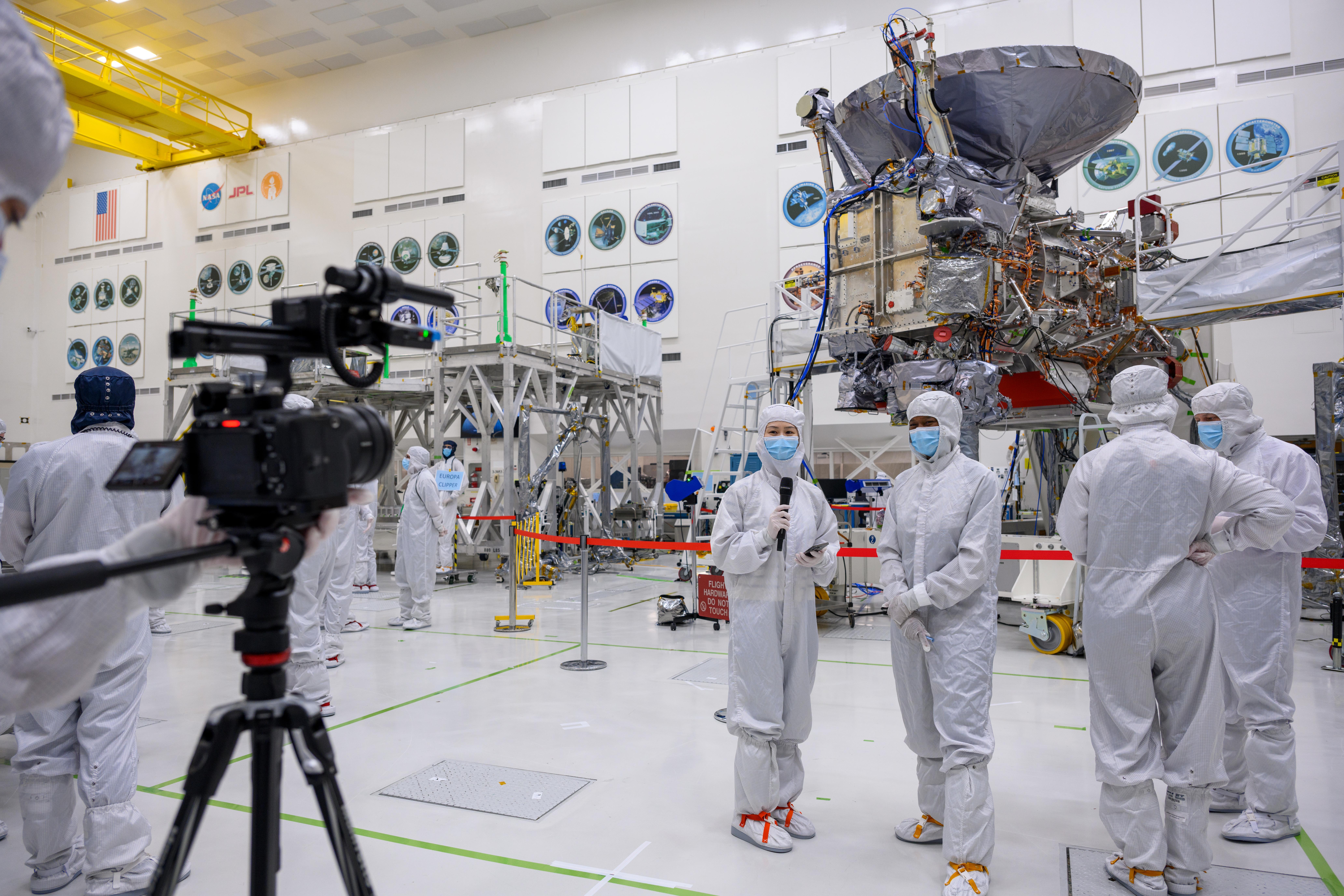 Journalists and workers in white protective clothing and blue face masks stand on a white floor near Europa Clipper. The spacecraft is behind them. It's mostly silver with lots of wires and cables exposed. A person with a video camera is in the bottom left of this image.