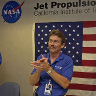 Rick Grammier clasping his hands together in front of a U.S. flag and the JPL sign.