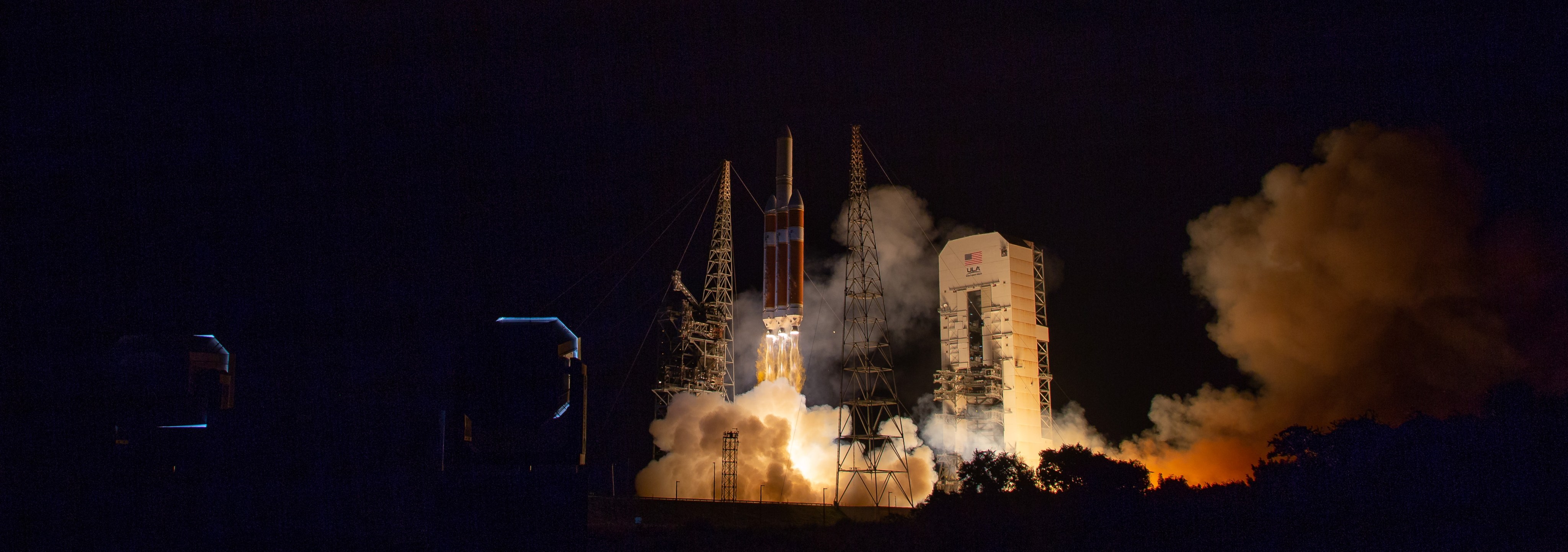 A rocket with orange and white boosters launches into the night sky. Clouds of smoke are beneath the rocket as it lifts off. A white launch tower lit with bright lights is nearby. Metal support towers also are visible.