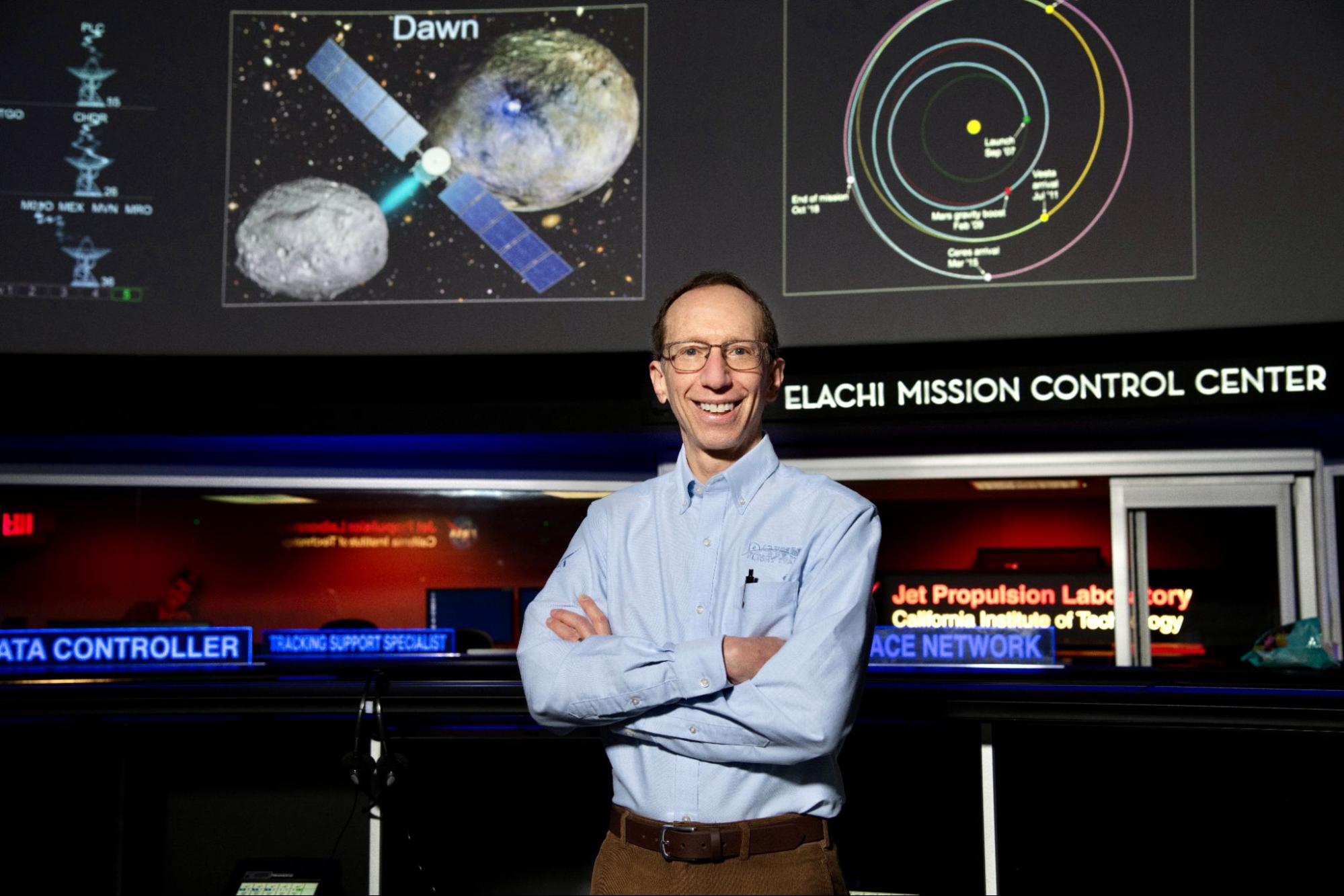 Man smiling with his arms crossed in front of a mission control panel