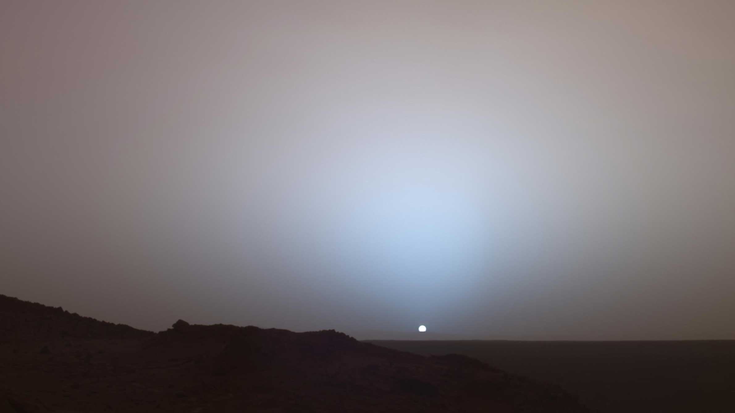 The Sun glows as it sets in the sky over Mars.