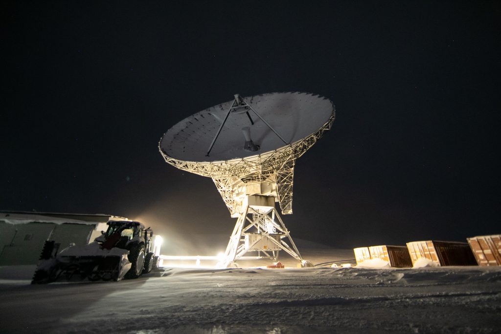 A large satellite dish is illuminated at night, surrounded by snow and some nearby structures and containers. A vehicle, possibly a snow plow, is parked to the left of the dish under a dark sky.