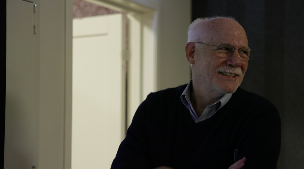 An older man with white hair and glasses, wearing a dark sweater over a collared shirt, stands smiling with his arms crossed in a dimly lit room near an open door.